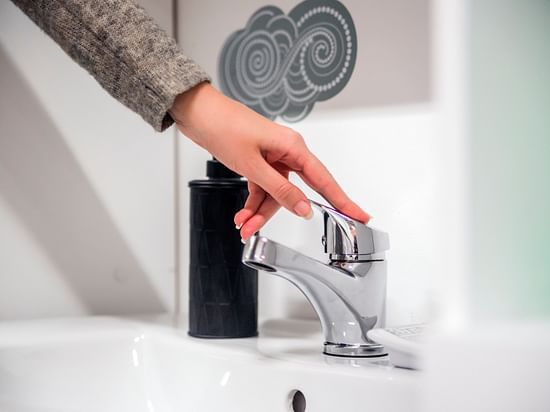 A hand turns on a modern chrome tap over a white sink in a bathroom setting.