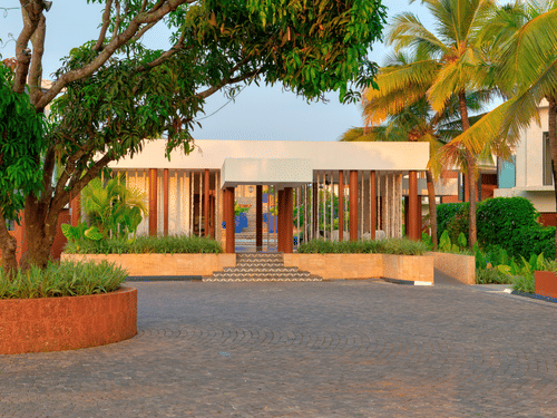 A modern white villa at Azaya Beach Resort Goa with timber pillars, surrounded by lush green trees and palms under a clear sky.