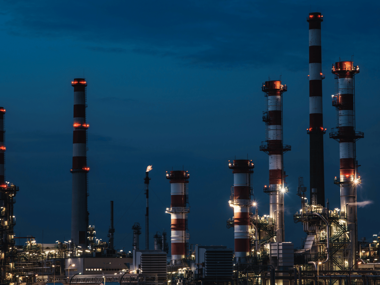 A night photograph of a large industrial oil refinery or chemical plant with tall, striped smokestacks illuminated in red and white against a dark blue sky.