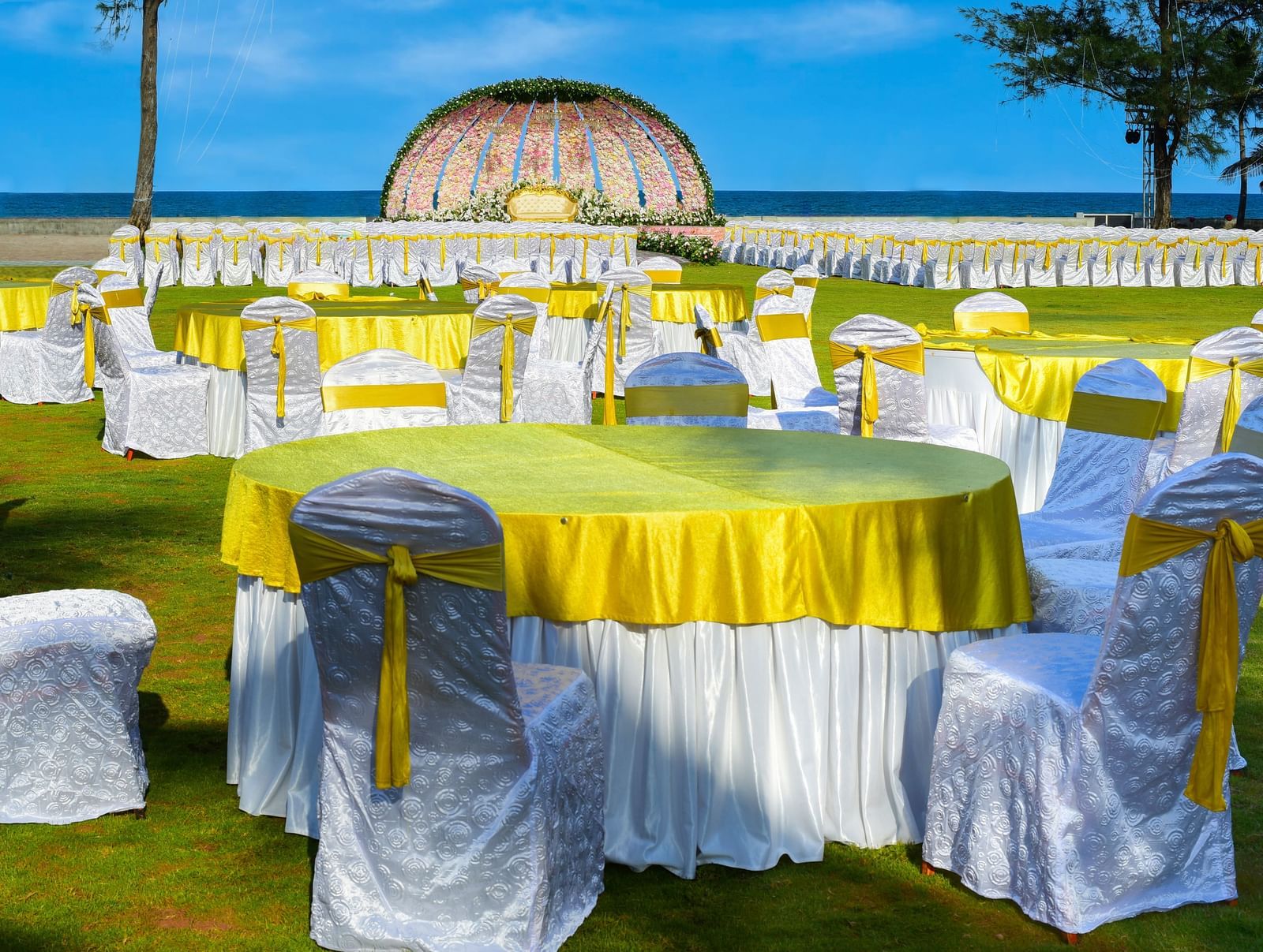 An outdoor event setup on a green lawn with yellow-draped round tables and white chairs, near a decorative archway and the ocean | Horizon | Grand Bay Resort and Spa