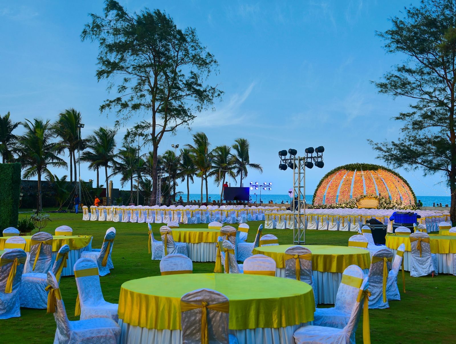 An outdoor evening event setup on a green lawn with round tables draped in yellow, featuring a lit stage and palm trees against a blue sky | Grand Bay Resort and Spa