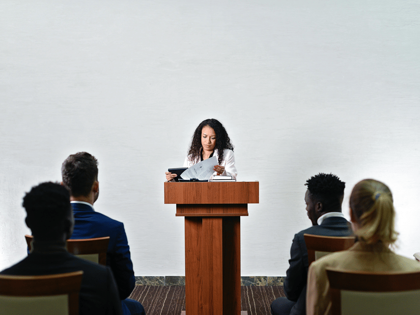 A woman standing behind a wooden podium, reviewing documents while four people sit facing her, listening attentively. The background is a plain white wall, and the audience members are dressed in formal attire.