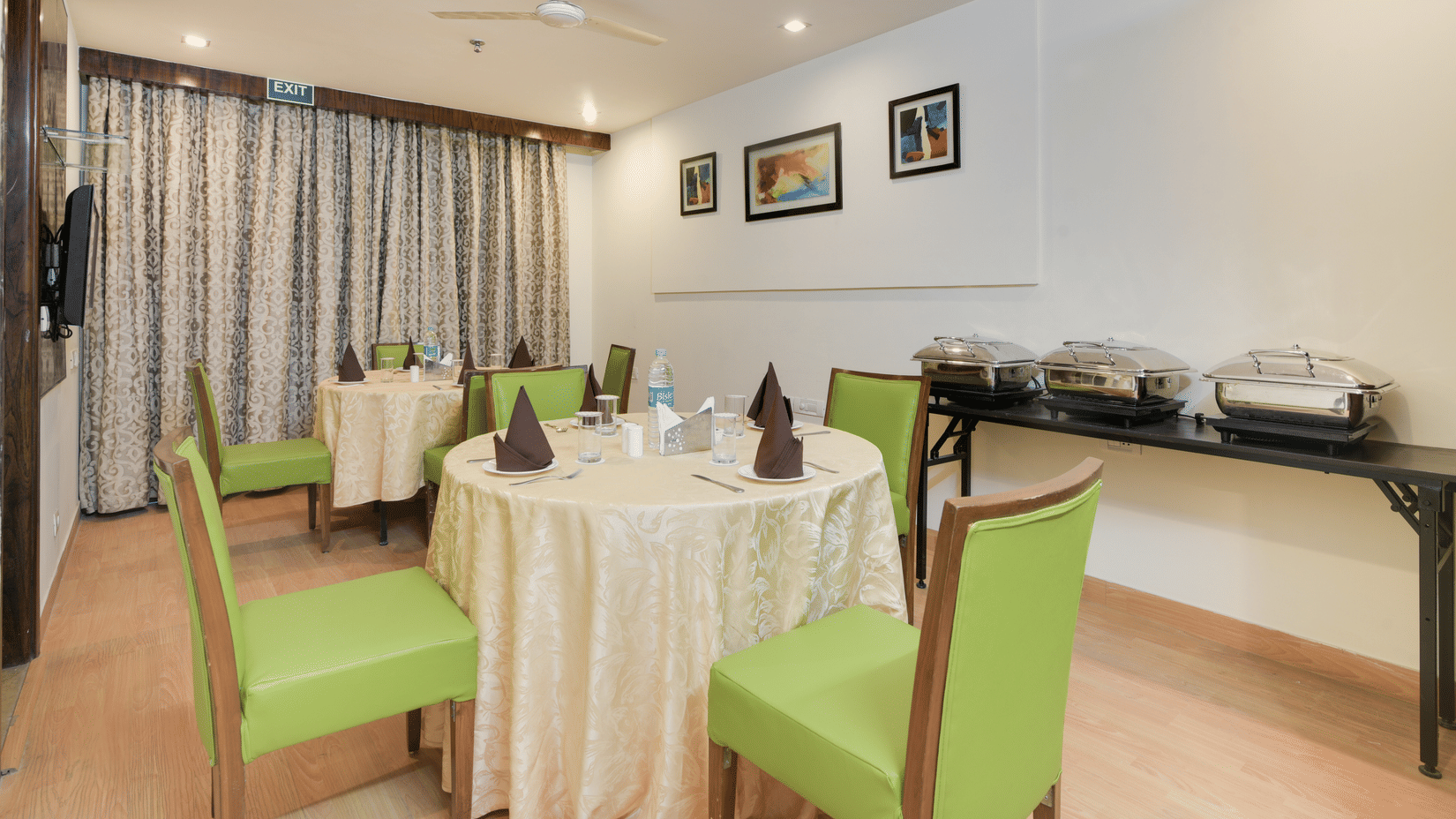 A small, brightly lit dining area at Hotel Bluestone Prime (Nehru Place), New Delhi featuring round tables with light green upholstered chairs and a buffet counter.