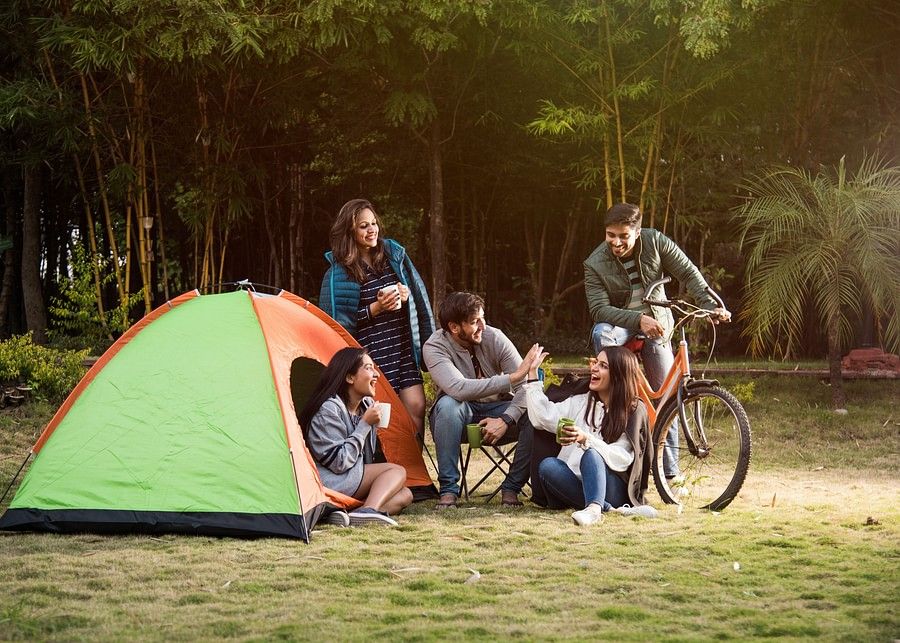  A group of six people, including adults and teenagers, are gathered around a campsite with a green and orange tent. They are enjoying a meal and drinks together, with bicycles parked nearby.