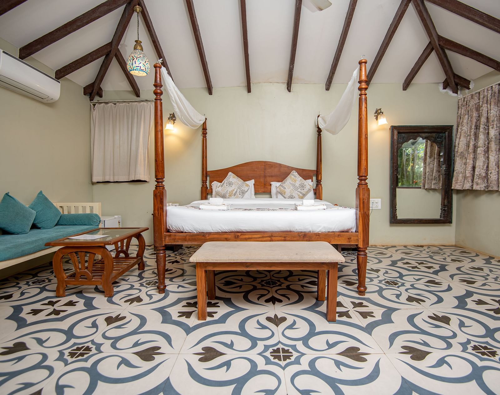 Spacious room with a large four-poster bed, cushioned sofa and wooden table, and a side window with curtains pulled aside at De Soul Santé, Morjim.