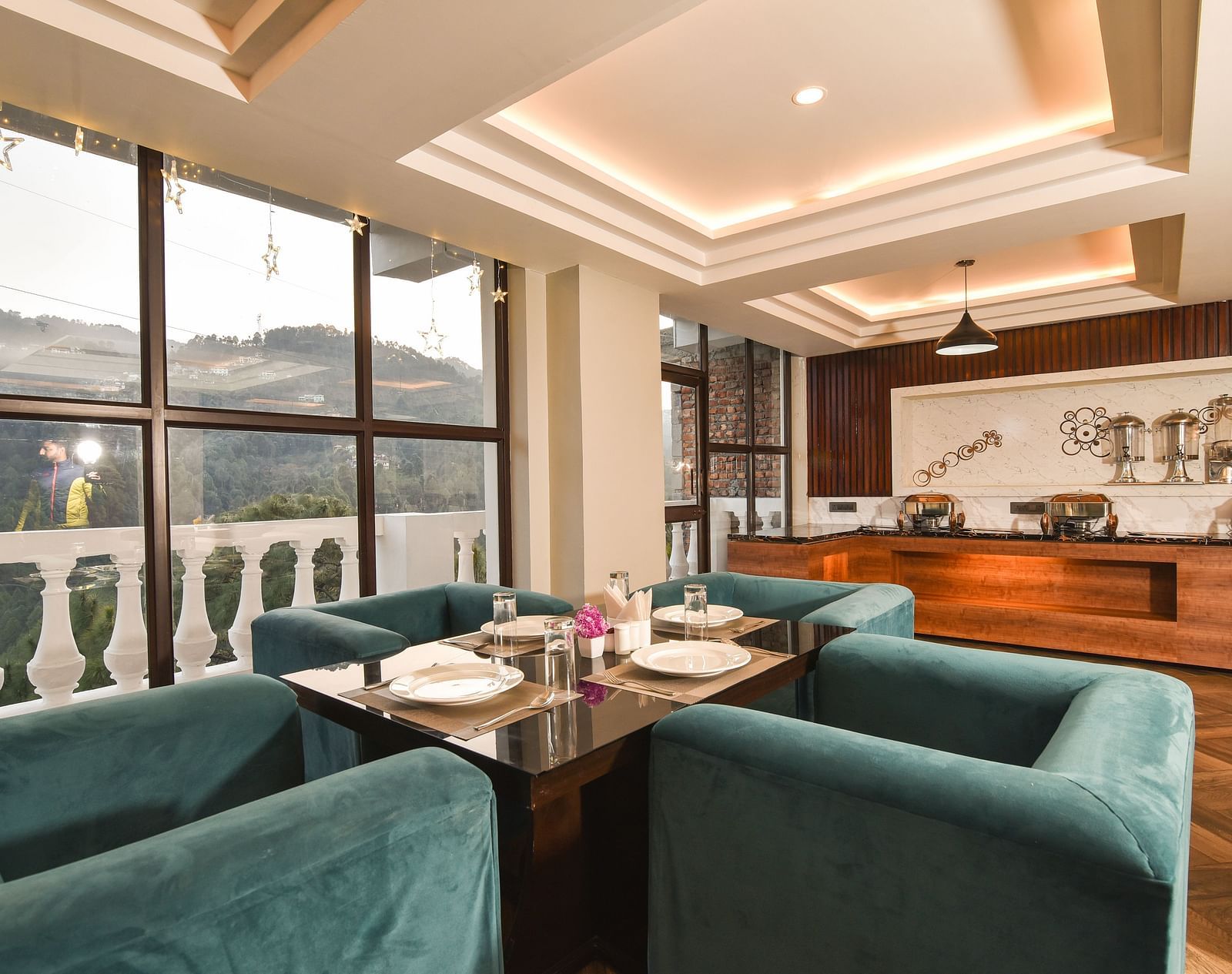 A modern dining area with teal upholstered chairs and a table set for two, with large windows offering scenic views of the mountains and landscape.