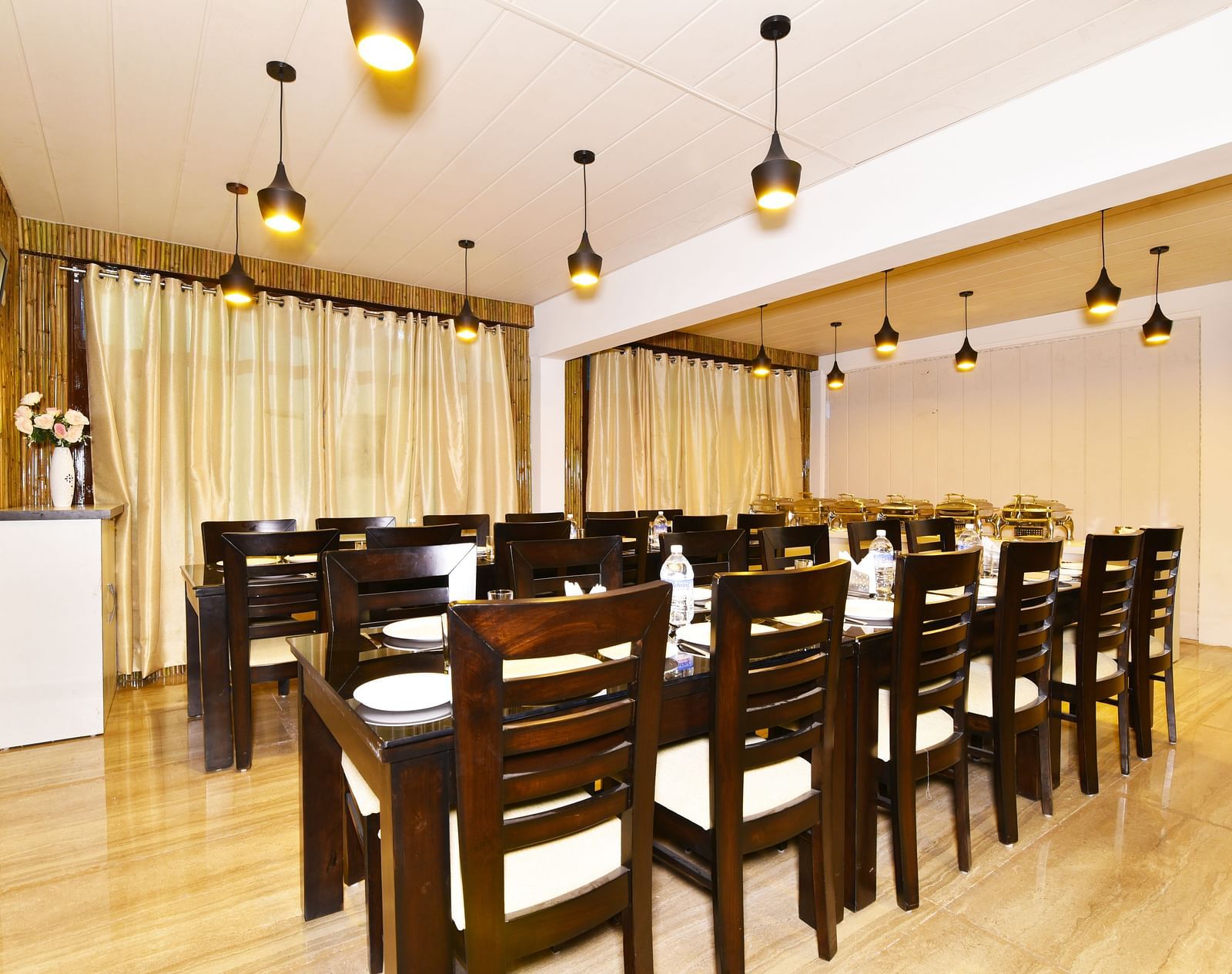 The image shows a spacious dining room with multiple tables and chairs, a buffet counter, and a large window with curtains | DLS Kapoor Resort Manali