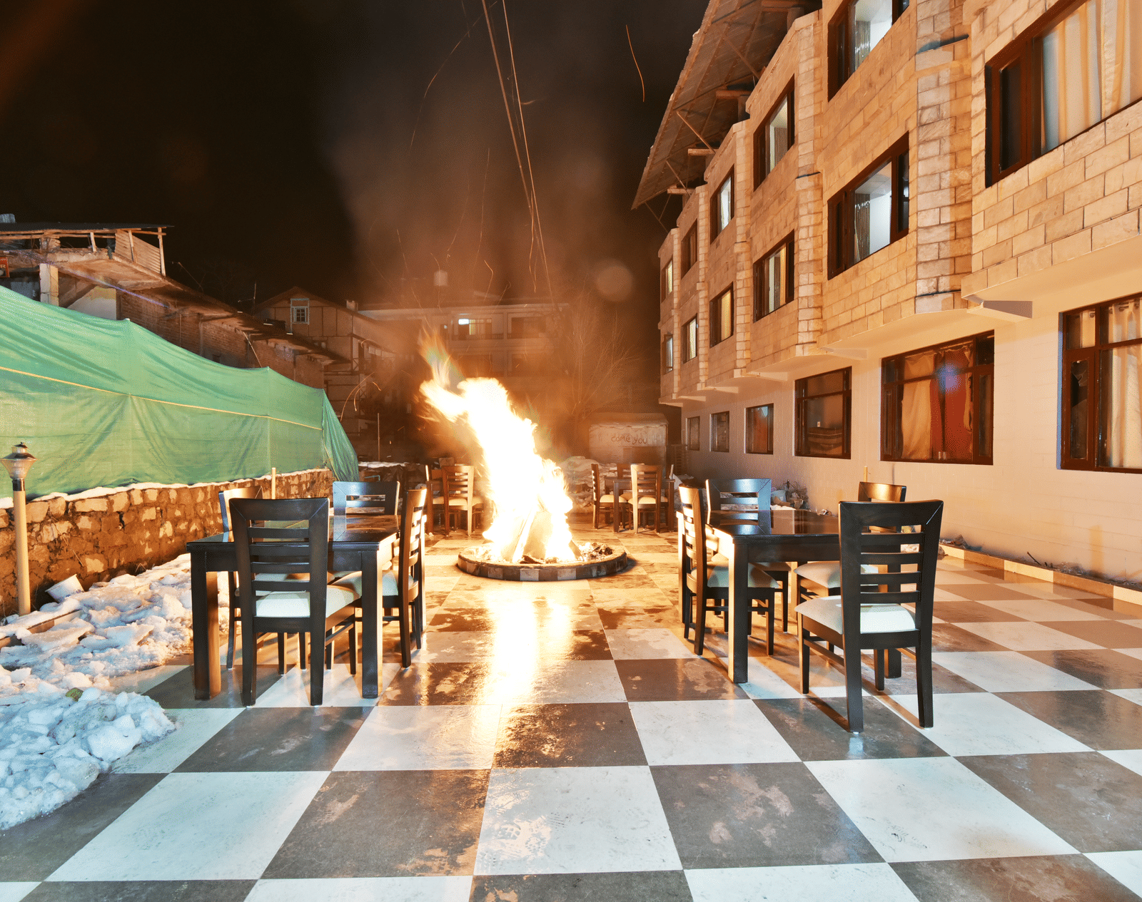 An overview of the outdoor dining area at night with a large bonfire in the centre, surrounded by tables and chairs - DLS Kapoor Resort, Manali.