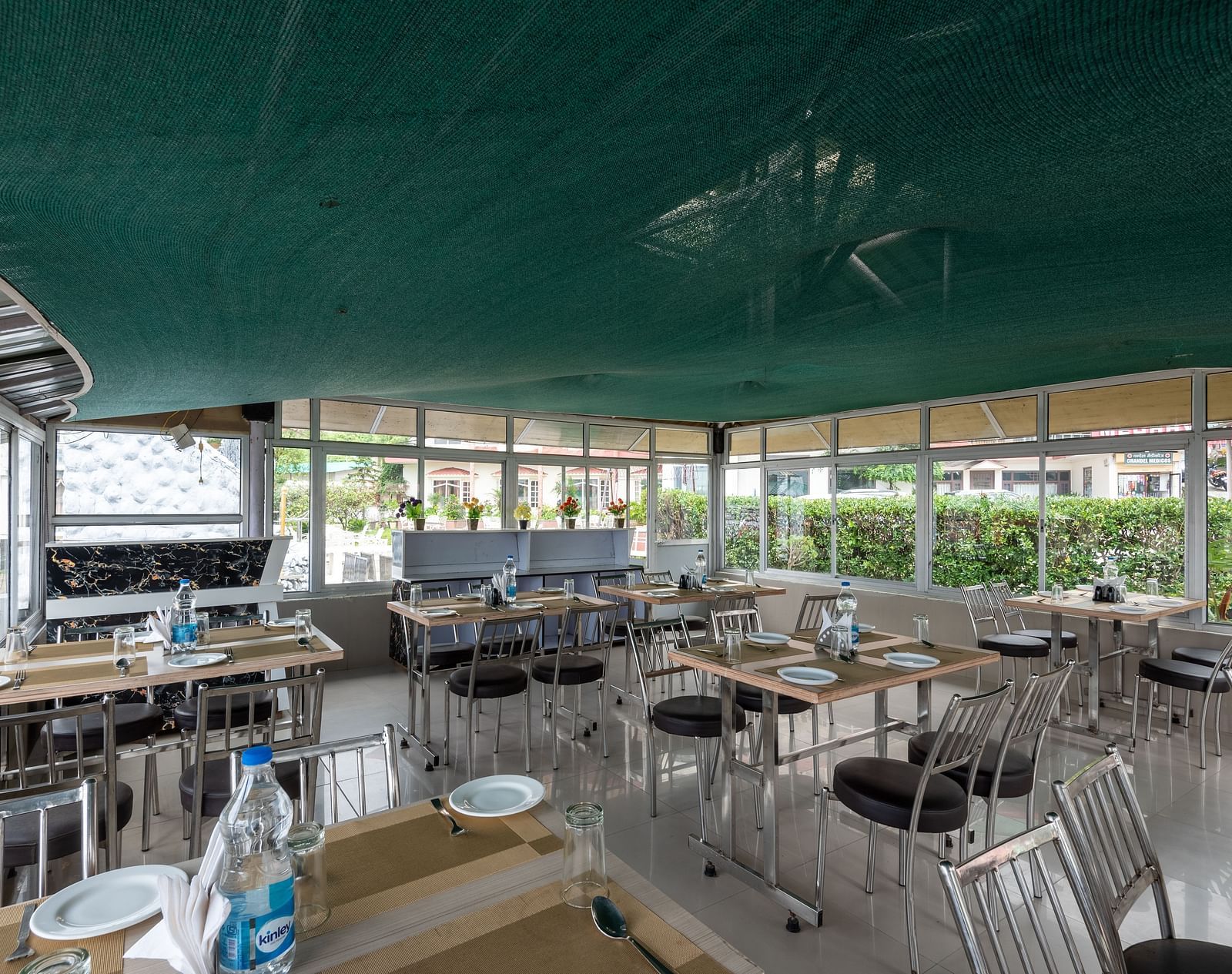 A cafe in Bhagsu with tables and chairs, featuring a green canopy overhead and large windows with views of the outside. The tables are set with plates, glasses, and bottles of water - DLS Meghavan Resort, Dharamshala