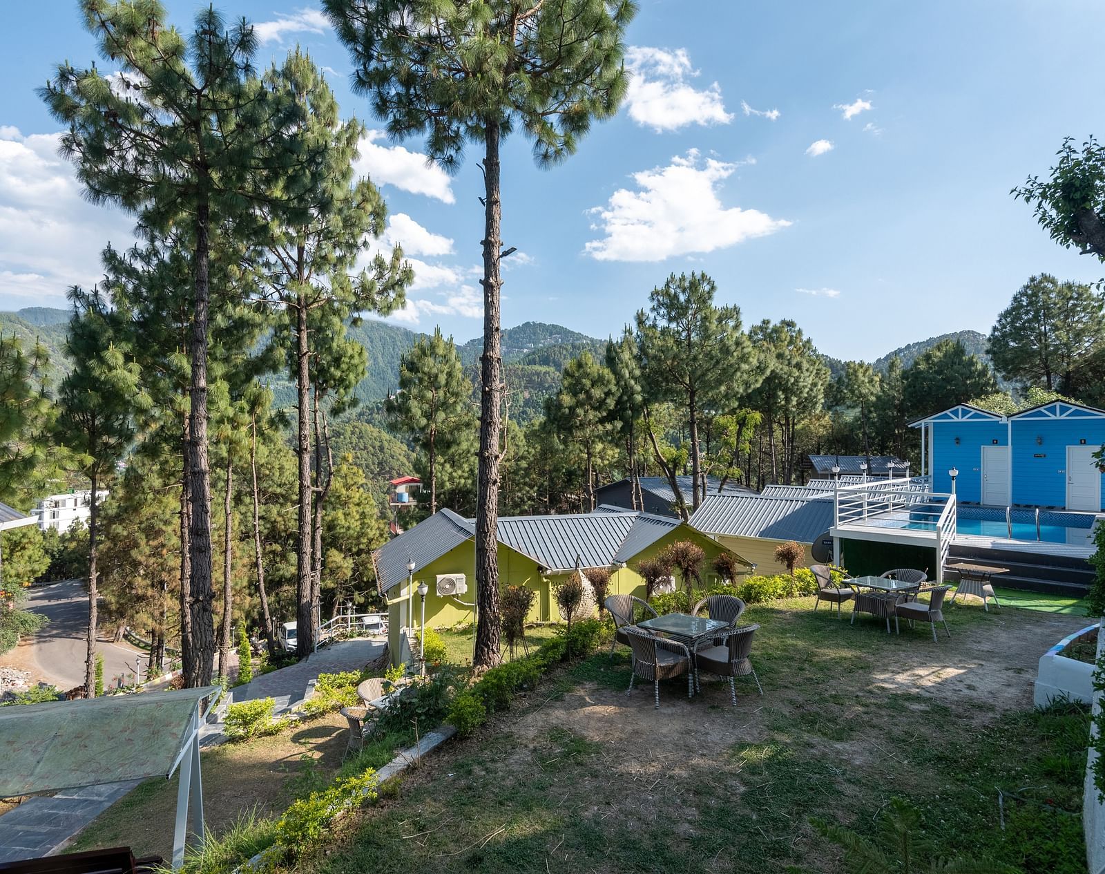 Scenic hillside resort with pine trees, outdoor seating area, and blue-roofed cottages set against a backdrop of lush green mountains under a clear sky - DLS Nature Trinket Resort, Dalhousie.