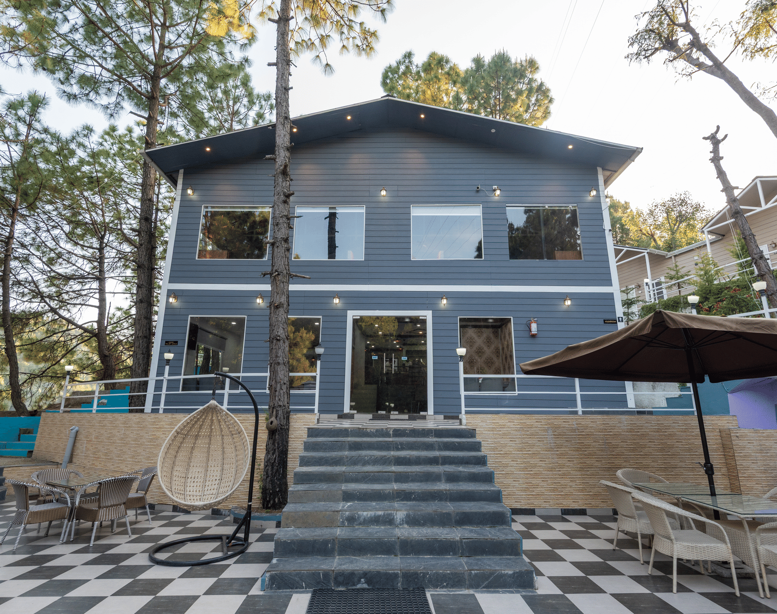 A modern two-story building with a dark blue facade and large windows, set against a backdrop of tall pine trees - DLS Nature Trinket Resort, Dalhousie, one of the best Hotels in Dalhousie Near Gandhi Chowk