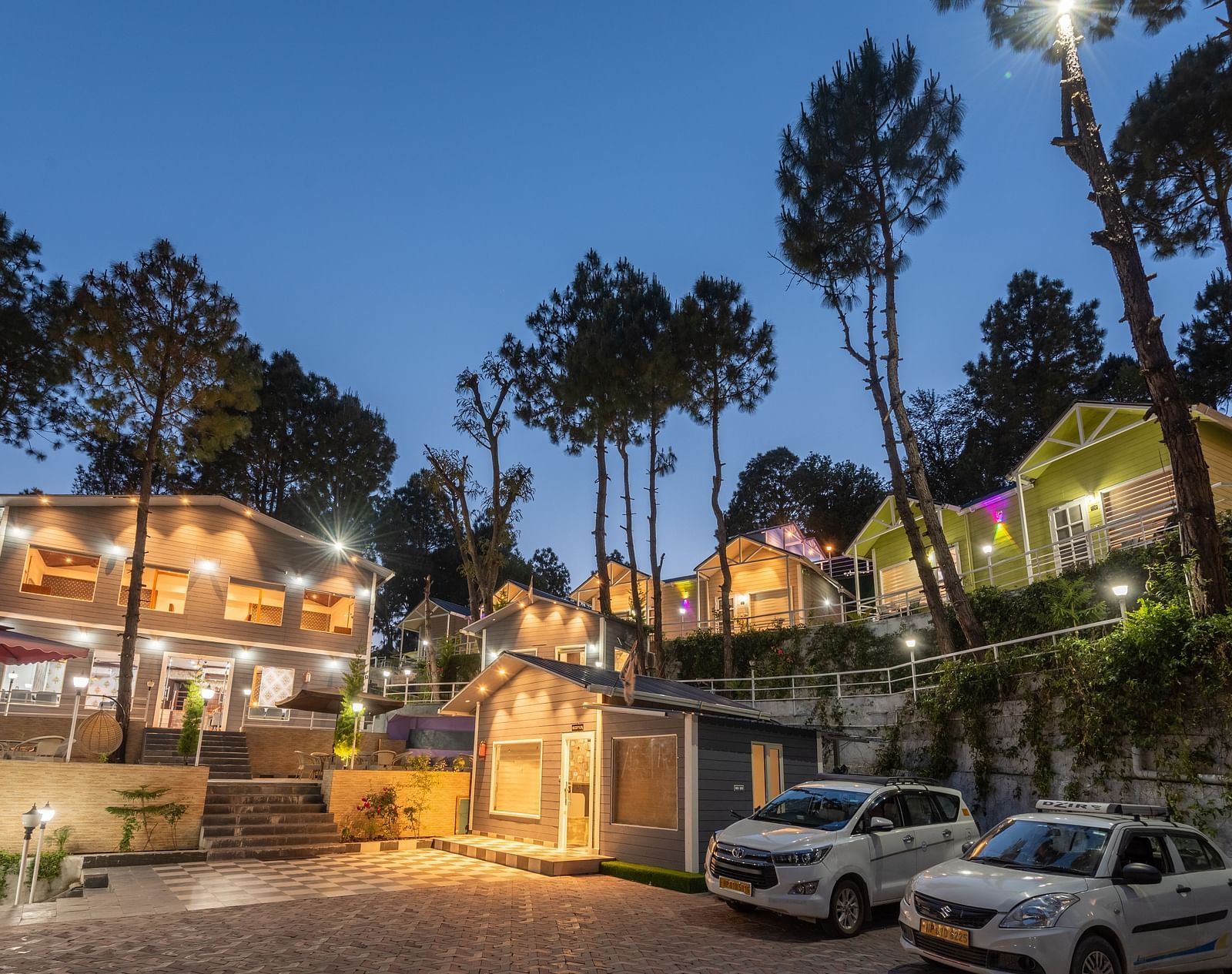 Twilight view of a hillside resort with warmly lit buildings, parked cars, and tall pine trees silhouetted against the evening sky - DLS Nature Trinket Resort, Dalhousie.