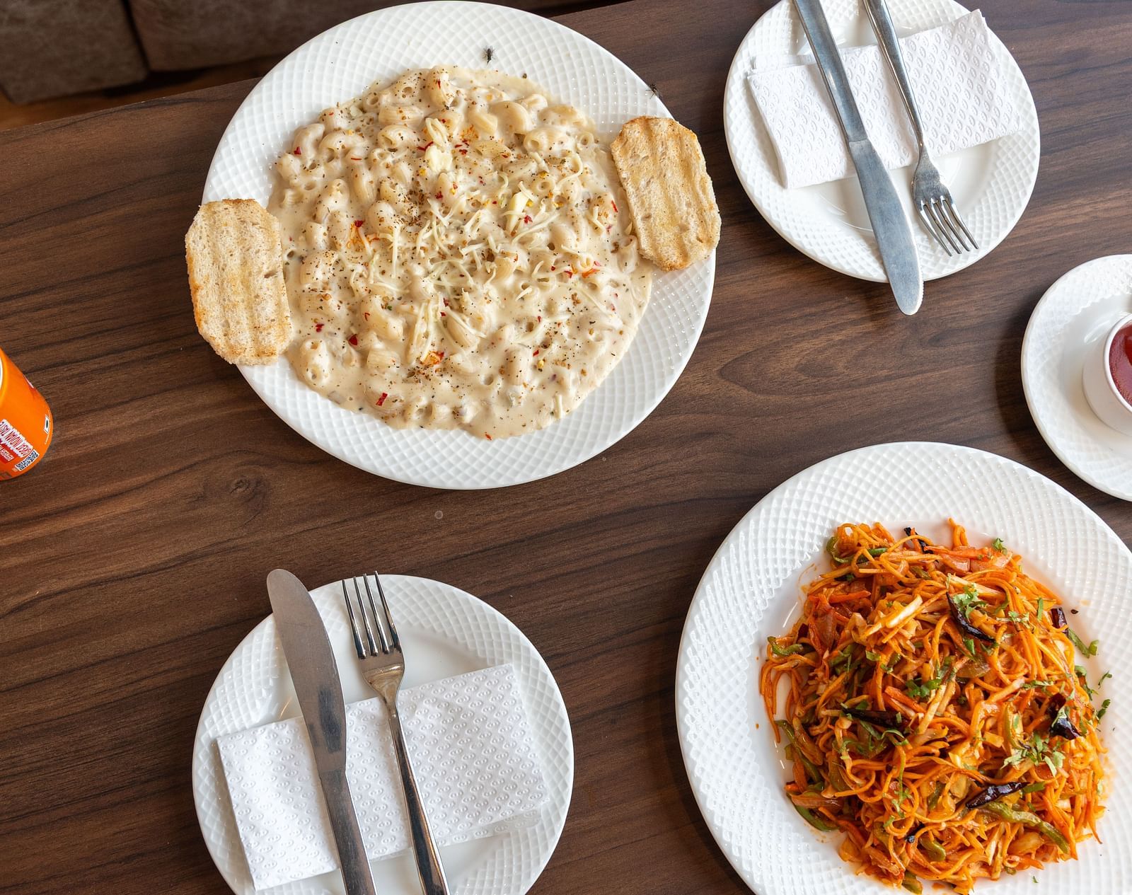 A delicious spread featuring a plate of creamy pasta with crispy breadsticks, alongside a plate of colourful noodles, and a serving of ketchup, complemented by a refreshing orange drink - DLS Nature Trinket Resort, Dalhousie.