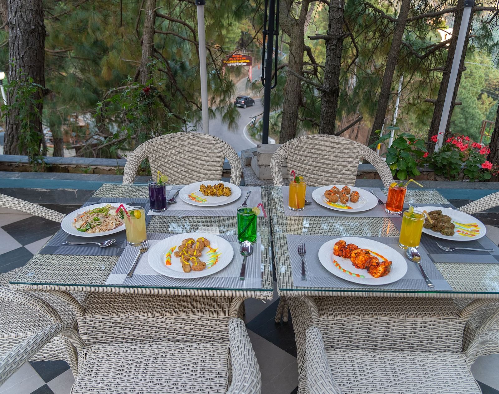 Outdoor dining setup with plates of food and drinks on a glass table, surrounded by wicker chairs and trees in the background - DLS Nature Trinket Resort, Dalhousie. 