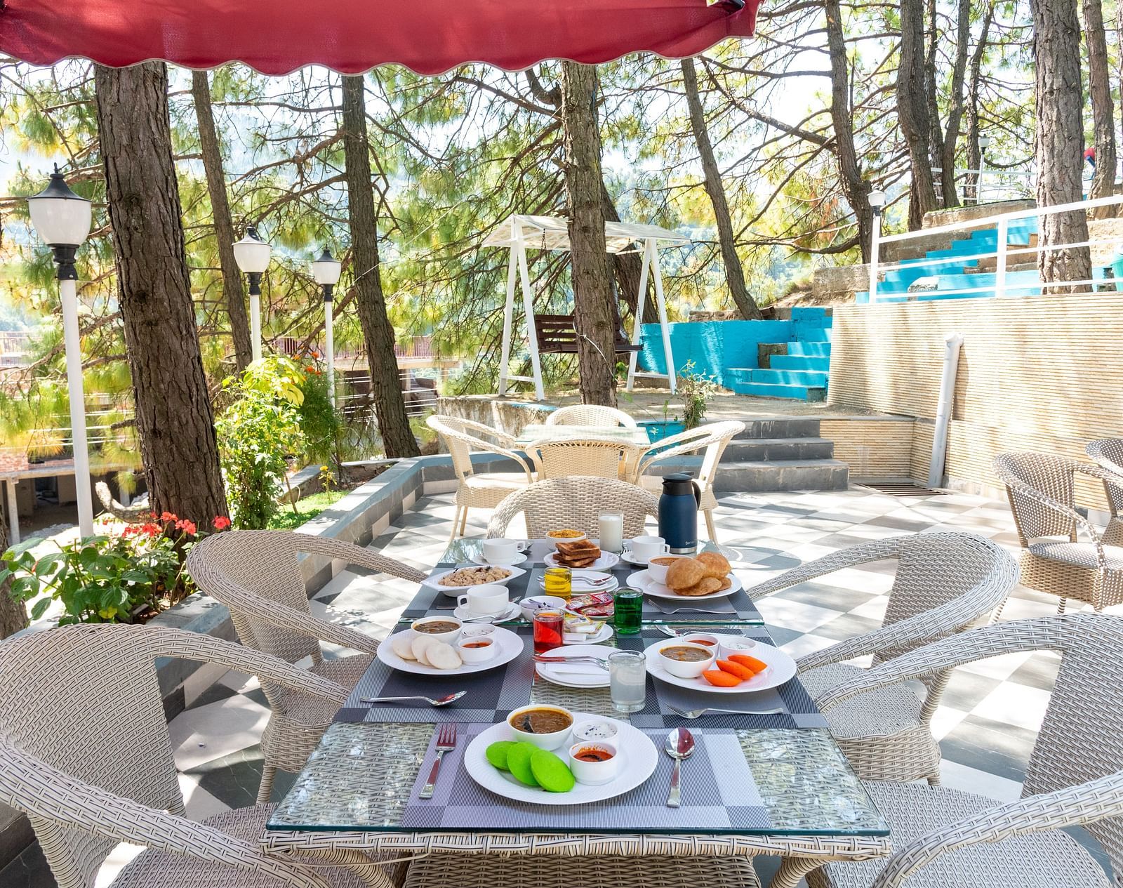 Spacious outdoor dining area set for breakfast, featuring a glass-top table with assorted dishes, surrounded by white chairs under a red canopy, nestled among tall trees and natural greenery - DLS Nature Trinket Resort, Dalhousie.