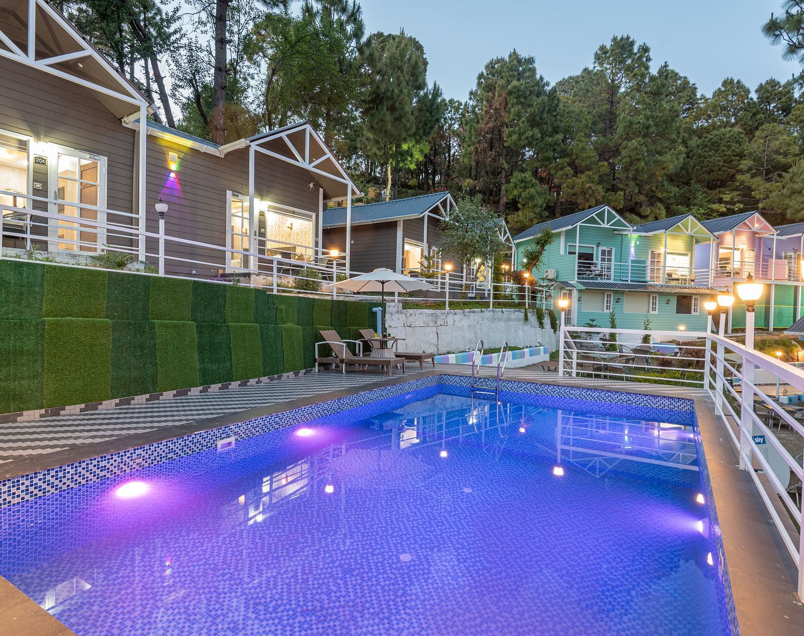 Evening view of a lit-up swimming pool with purple and white underwater lights, surrounded by a safety railing, colourful cottages, and pine trees in a hillside resort setting - DLS Nature Trinket Resort, Dalhousie.