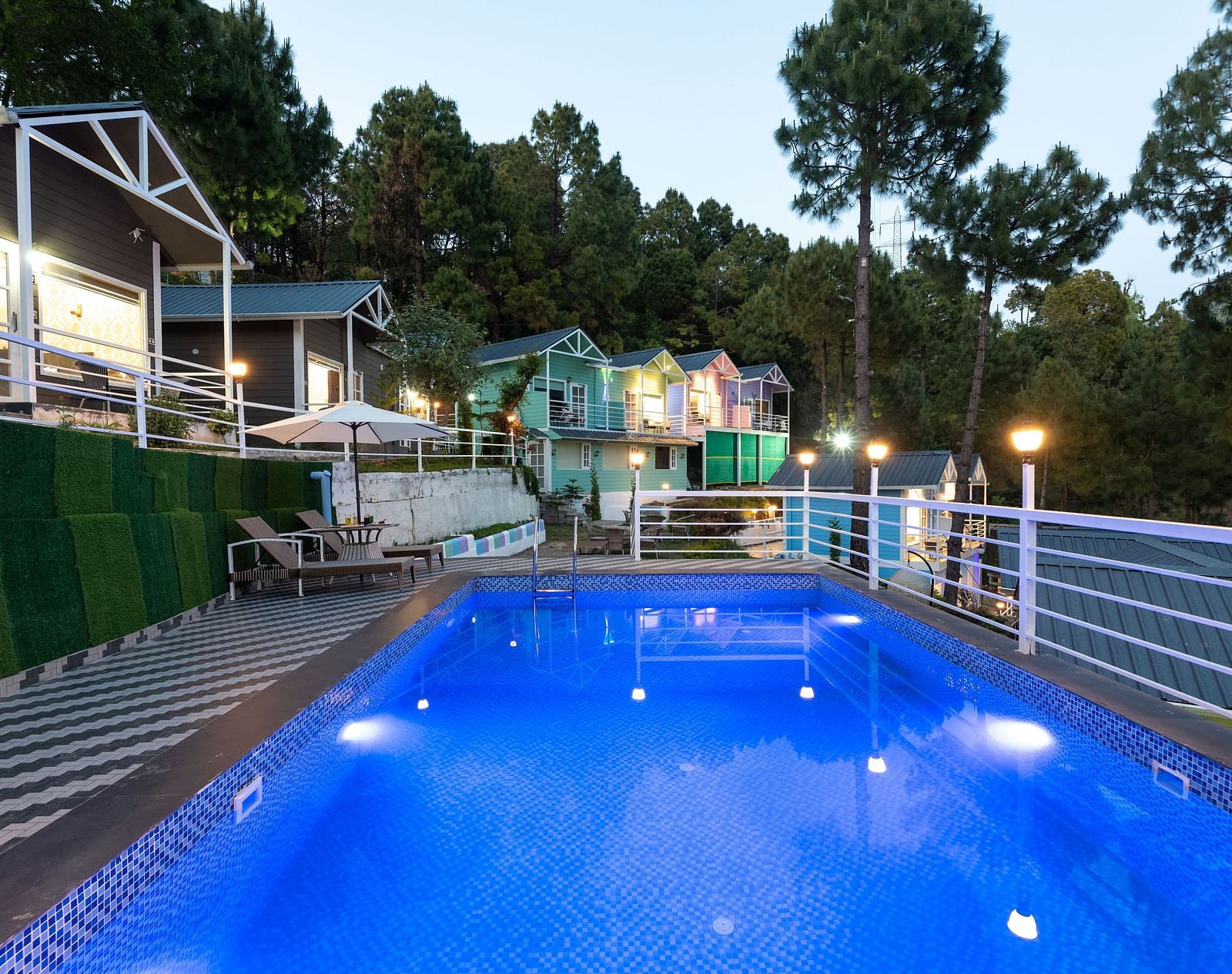 Tranquil evening view of a lit swimming pool surrounded by safety railings and striped flooring, with cosy hillside cottages and tall pine trees in the background - DLS Nature Trinket Resort, Dalhousie.