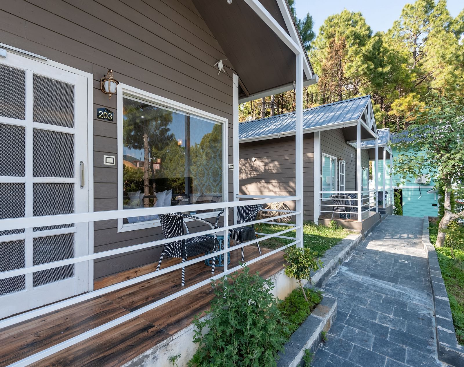 Row of modern wooden cottages with front patios, white railings, and a stone pathway surrounded by greenery and tall pine trees - DLS Nature Trinket Resort, Dalhousie.