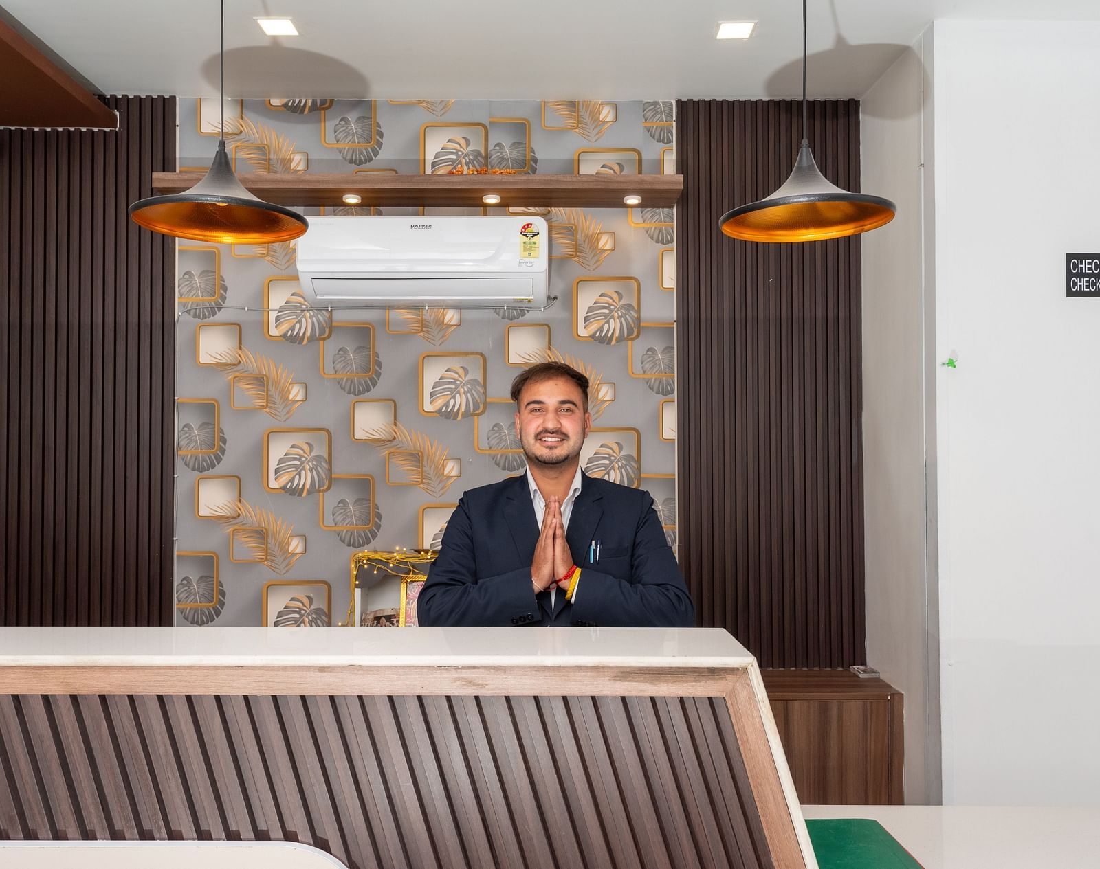  A smiling man in a dark suit stands behind a modern reception desk with folded hands in a greeting gesture, set against a backdrop of patterned wallpaper, pendant lights, and a wall-mounted air conditioner - DLS Nature Trinket Resort, Dalhousie.