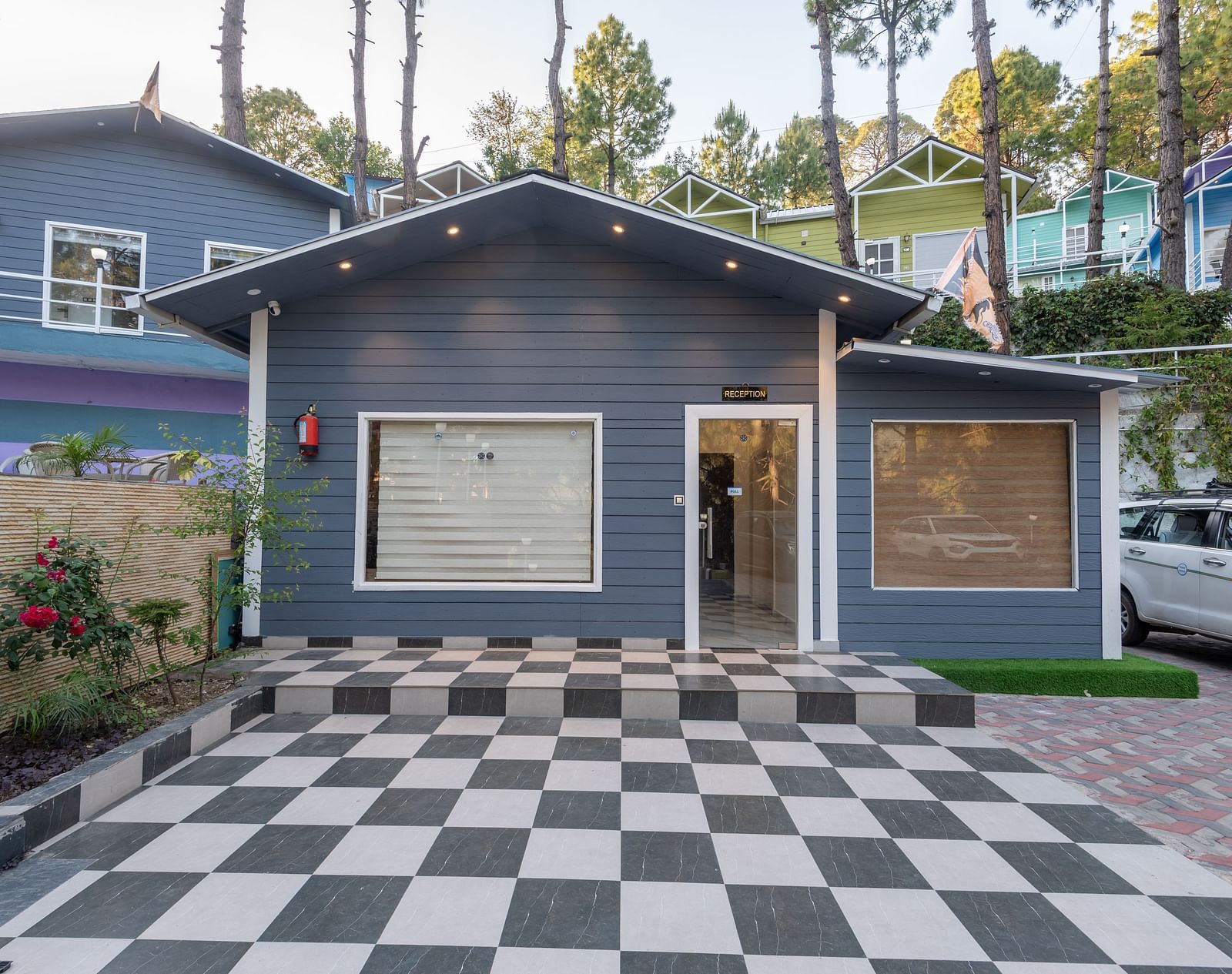 A modern, single-story building with dark grey siding, large front windows, and a checkered black-and-white tile driveway, surrounded by colourful neighbouring houses and trees in the background - DLS Nature Trinket Resort, Dalhousie.