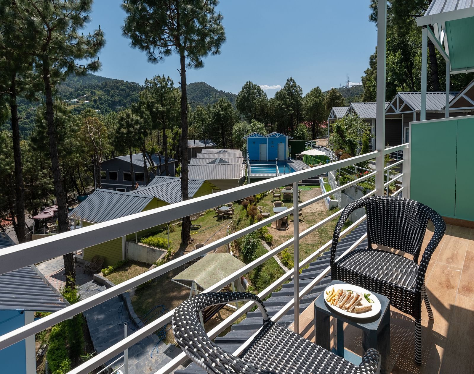 A balcony at DLS Nature Trinket Resort, Dalhousie, features two black wicker chairs and a small table with a plate of food, overlooking a scenic view of buildings, greenery, and tall trees under a clear sky.