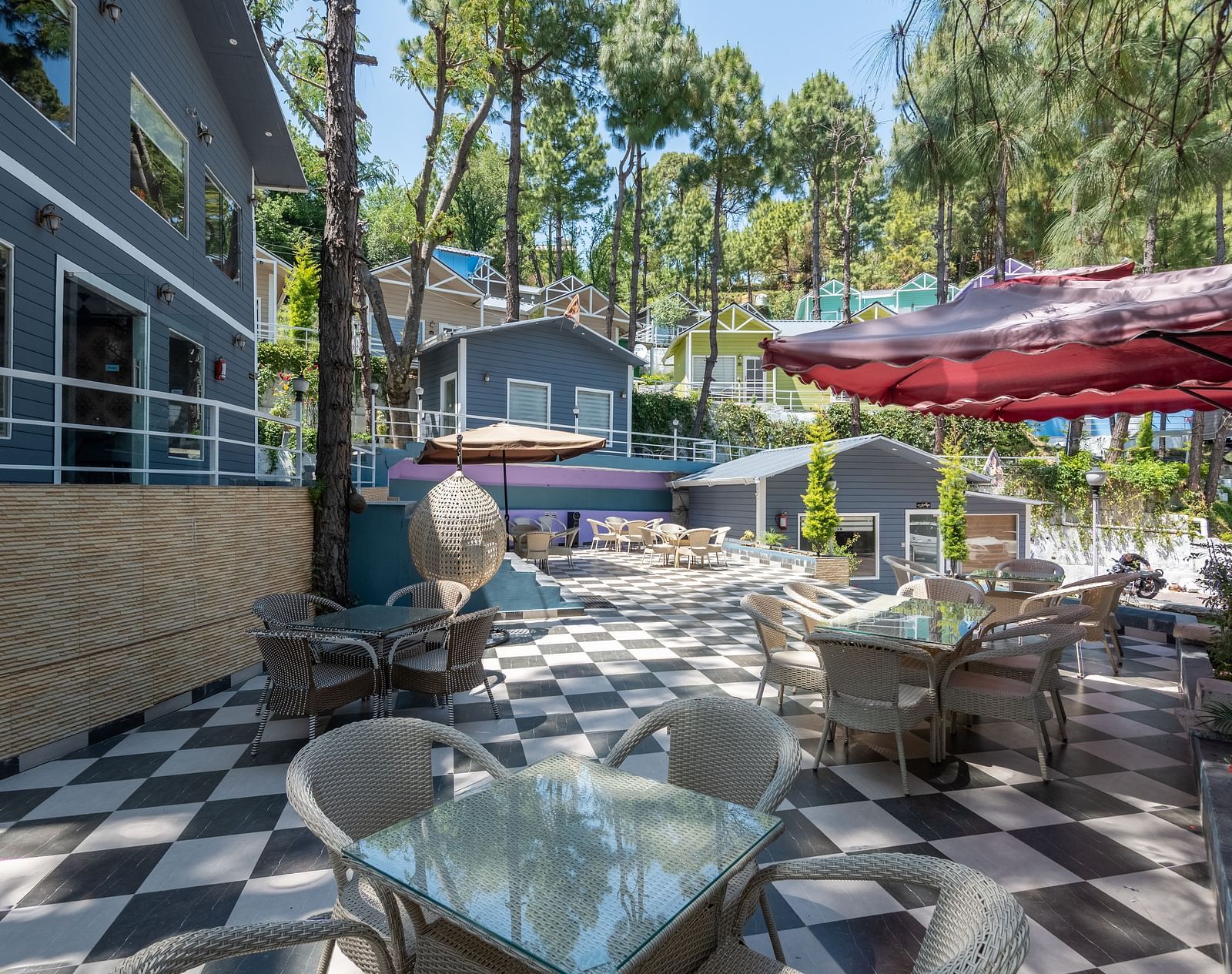 Sunny outdoor café area with glass tables, red umbrellas, and black-and-white checkered flooring, bordered by grey buildings and tall pine trees - DLS Nature Trinket Resort, Dalhousie.