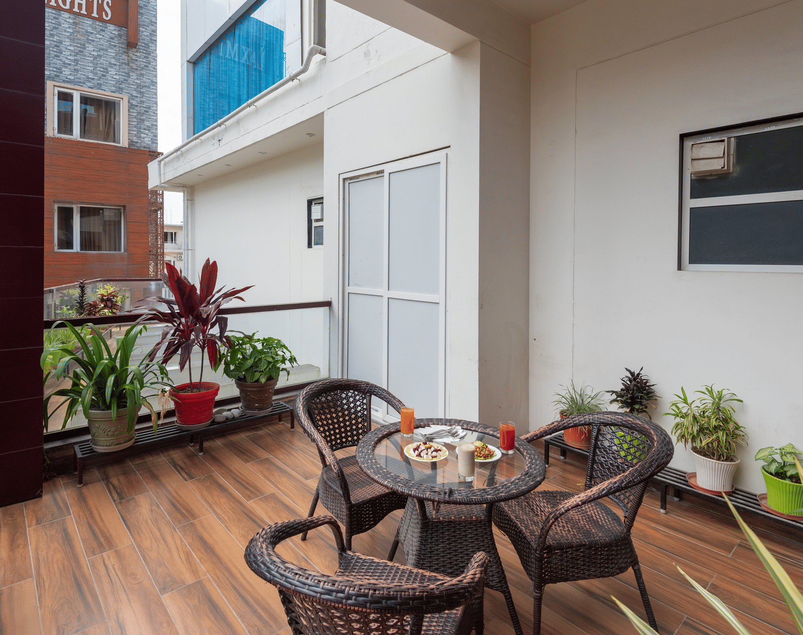 Alt text: A close-up of a balcony at DLS Hotels On The Ganges. A small, glass-topped table with a wicker base is surrounded by three matching chairs. The table is set with food and drinks. The balcony has a mix of wood-like tiled flooring and white walls