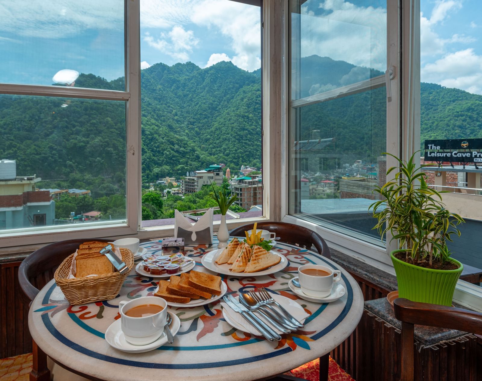 A cafe table is set for breakfast, with plates of toast, grilled sandwiches, jam, and coffee. The setting is bright with natural light from large windows, offering a view of a green, forested mountain and a townscape below. This is a shot from the cafe at 