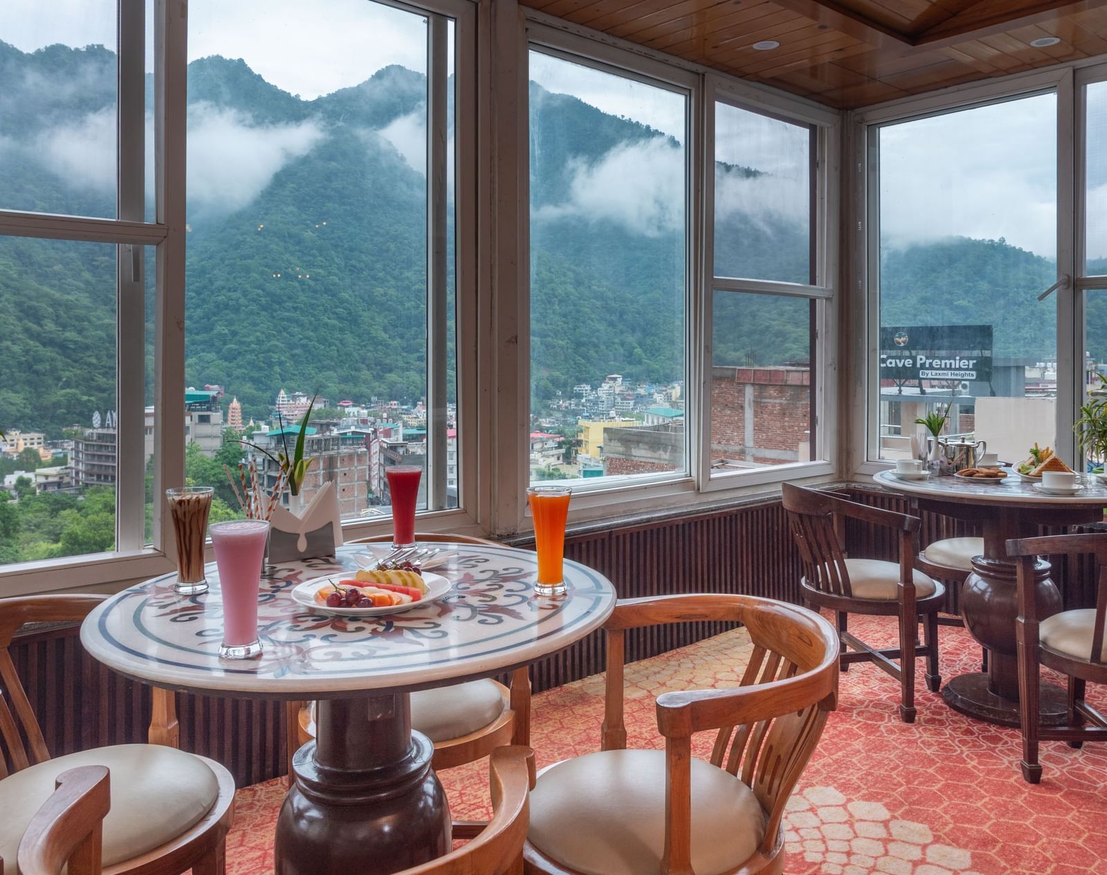 A cafe with a vibrant, red patterned carpet and wooden furniture. The room has large windows that look out onto misty green mountains and the city below. Tables are set with various drinks and food, including a fruit platter and sandwiches. This is the caf
