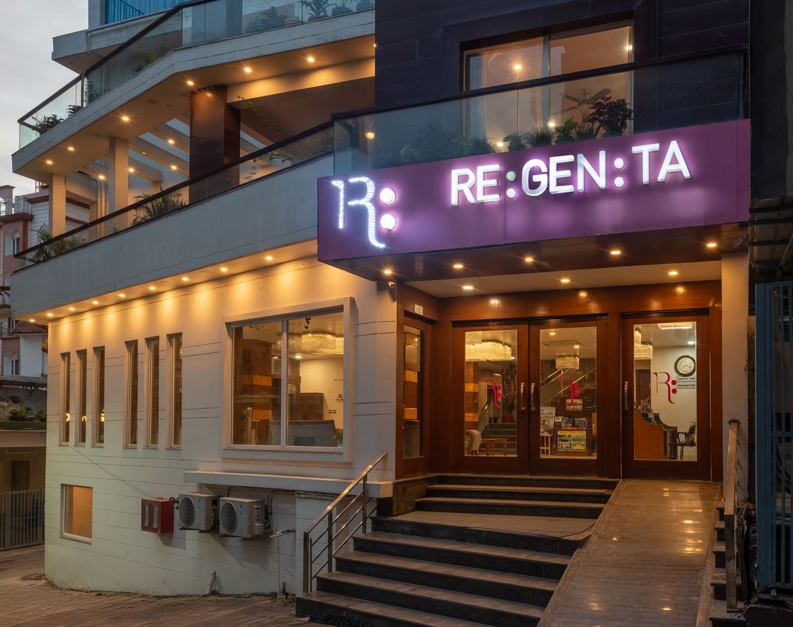 An eye-level view of the entrance to the DLS Hotels On The Ganges, in the evening. The hotel's brightly lit RE:GEN:TA sign is visible, along with the glass-paned doors of the lobby. The exterior walls are a mix of white and dark paneling, with warm light