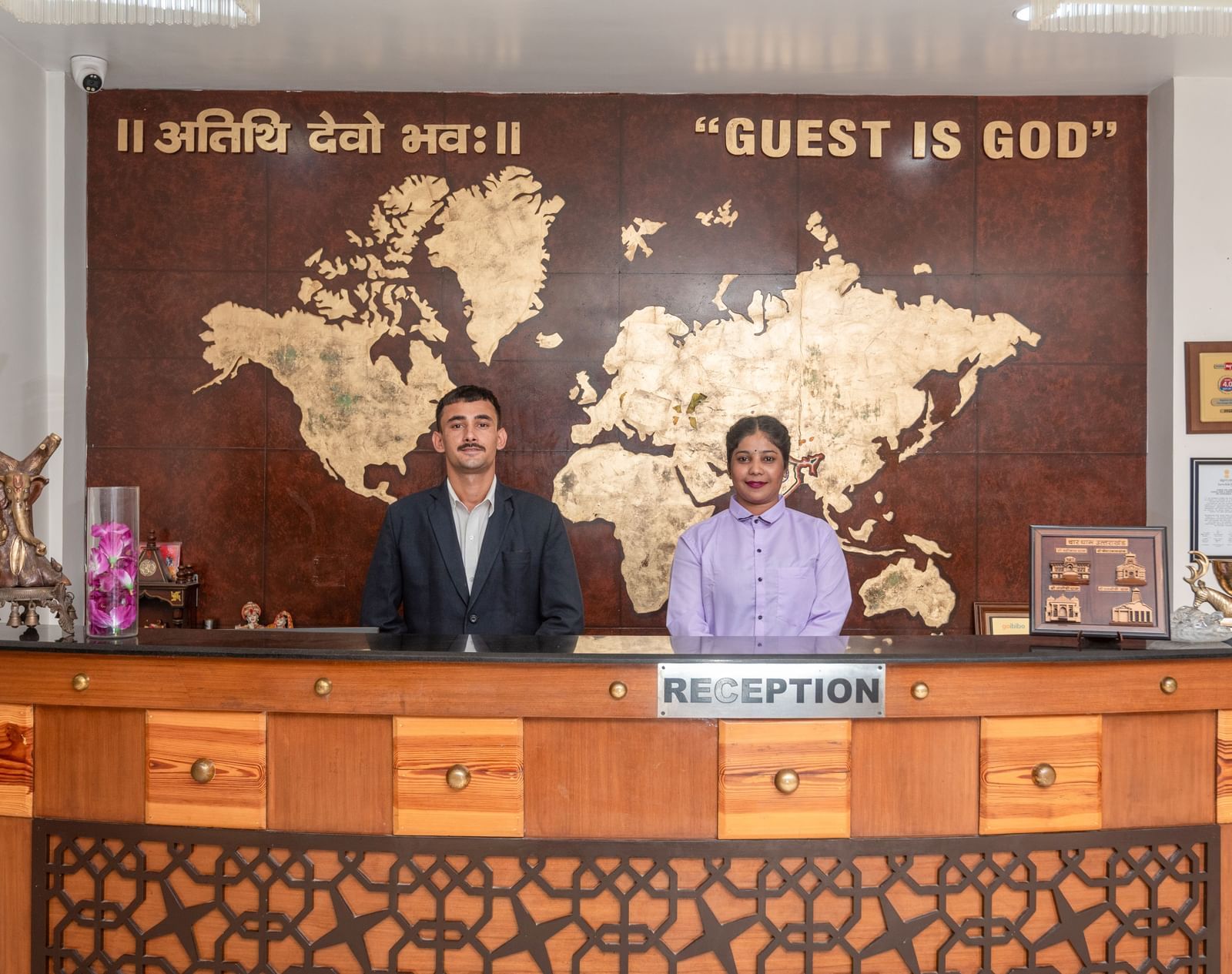 Two hotel staff members, a man and a woman, stand behind a wooden reception desk. Behind them is a large wall with a world map and the phrase Guest Is God in English and Hindi. This is the reception area at DLS Hotels On The Ganges.