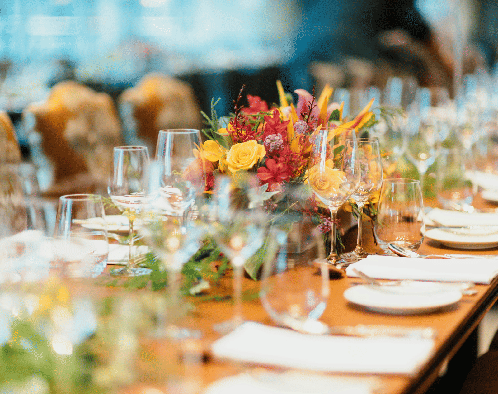 A shallow-focus shot of a long wooden dining table set with wine glasses and bright floral decorations.