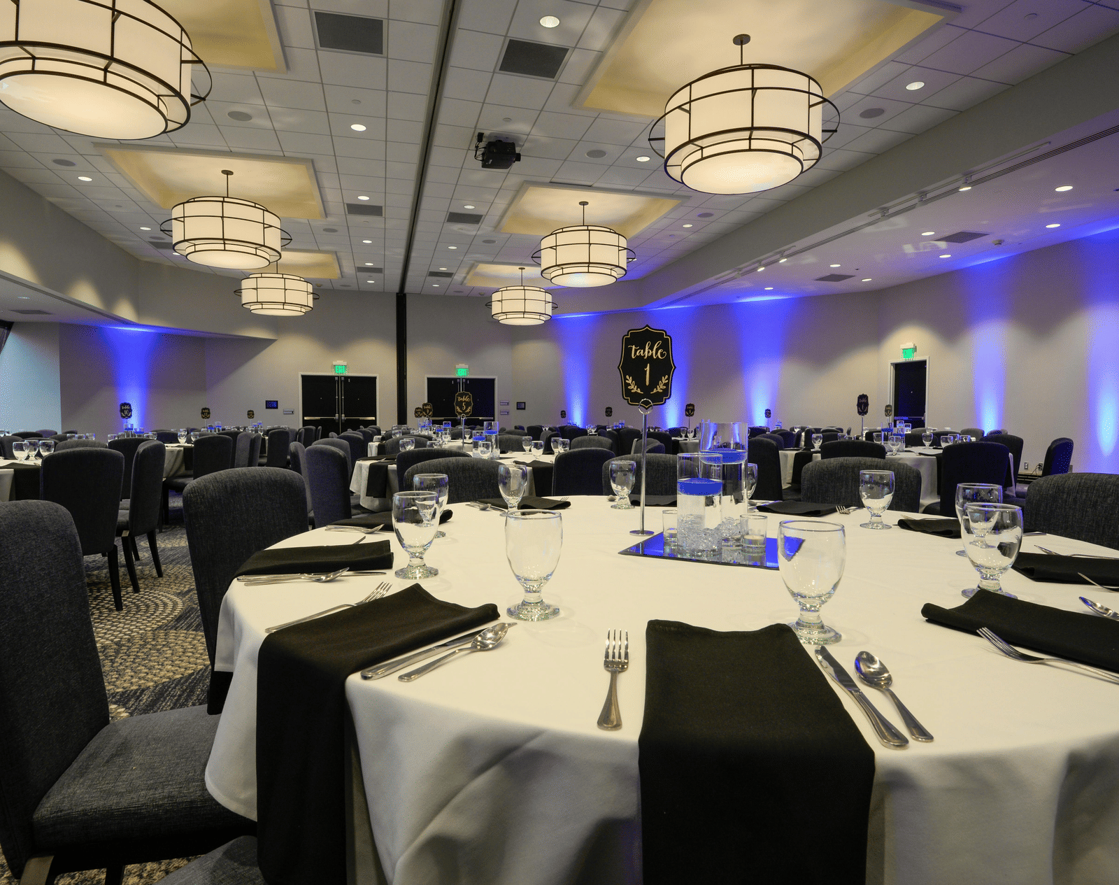 A wide view of a banquet hall set up with large, round tables, white linens, black napkins, and modern chandeliers. 