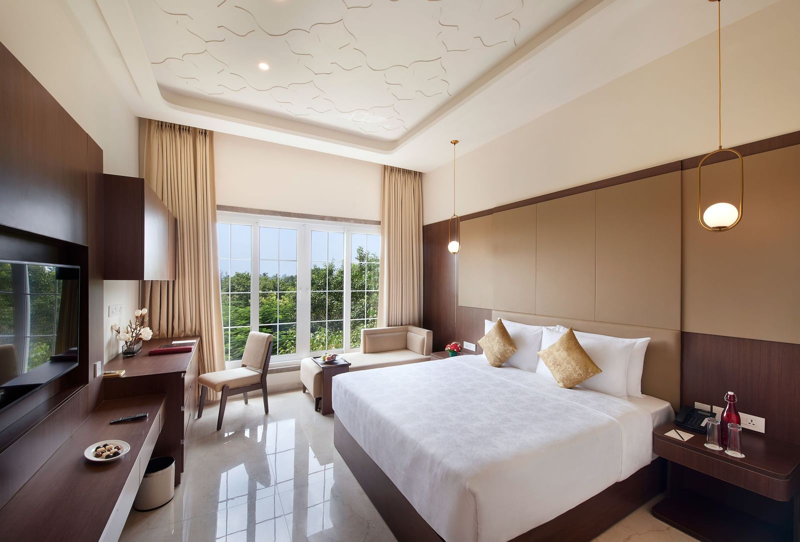 A hotel room in Chennai with a double bed, a wall-mounted television, hanging lamps, and a window offering views of greenery - Accord Chrome Airport Hotel