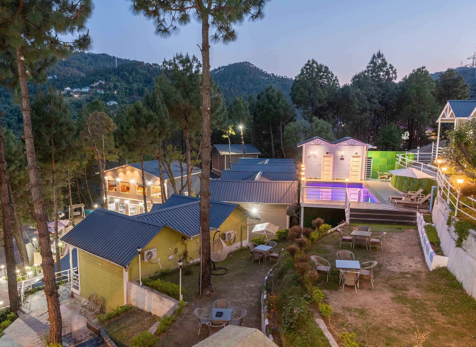 Elevated twilight view of a serene hillside resort with cosy cottages, an illuminated swimming pool, outdoor seating, and tall pine trees set against a backdrop of forested hill - DLS Nature Trinket Resort, Dalhousie.