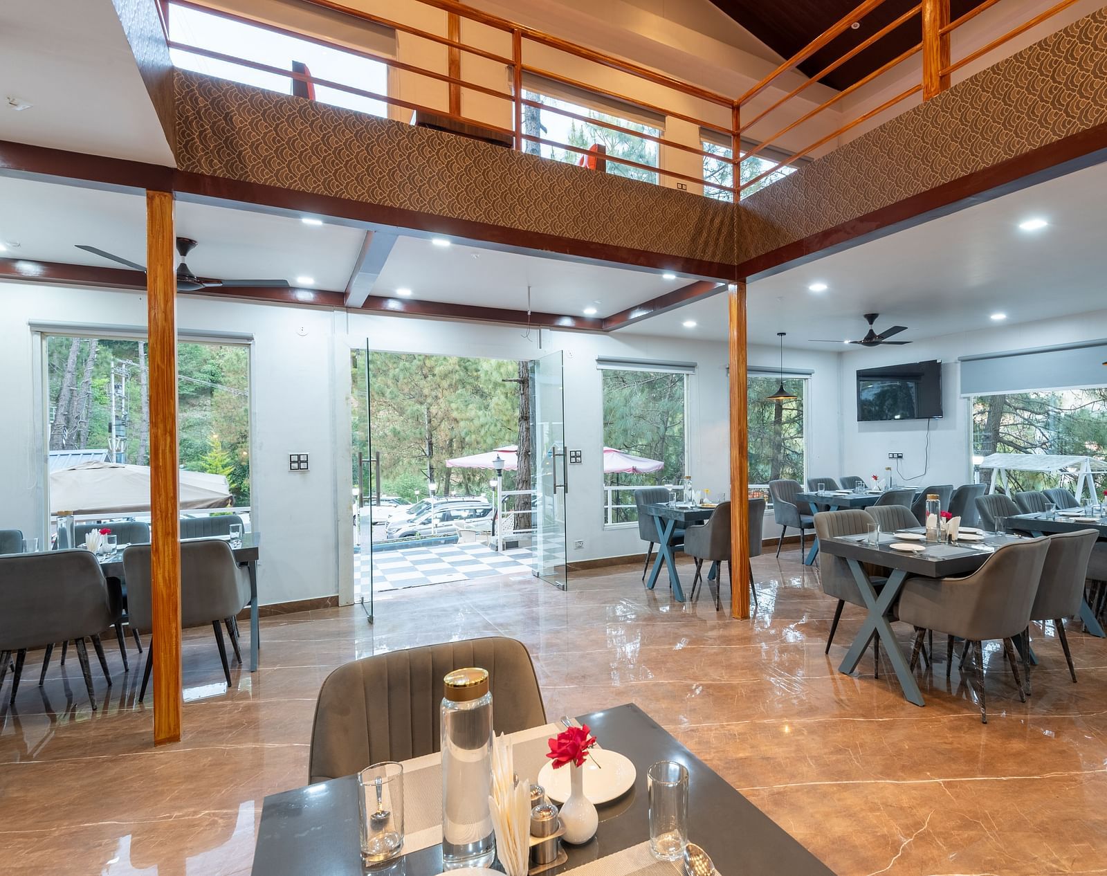 Bright and modern restaurant dining area with neatly arranged tables, grey chairs, large windows, and an open mezzanine level - DLS Nature Trinket Resort, Dalhousie.