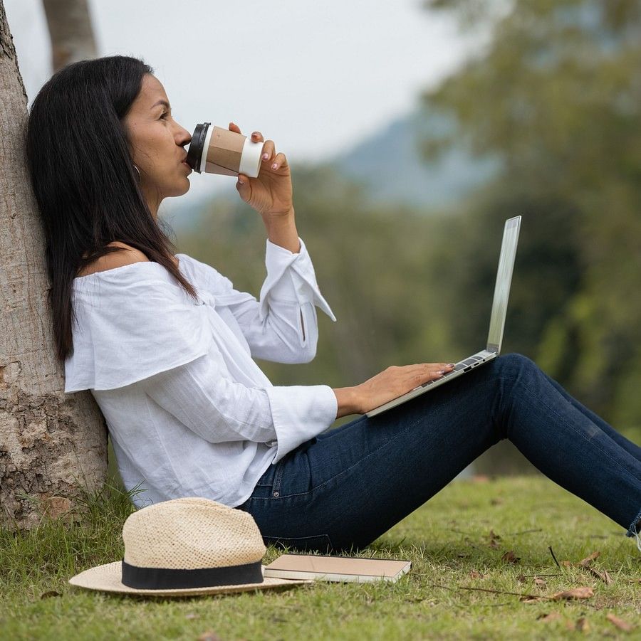 An image showing a woman drinking a cup of coffee, while sitting on the grass with her back on a tree, with a fedora hat and a laptop with her, and bushy landscapes in the backdrop