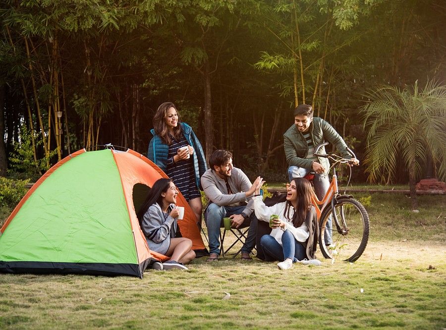  A group of six people, including adults and teenagers, are gathered around a campsite with a green and orange tent. They are enjoying a meal and drinks together, with bicycles parked nearby.