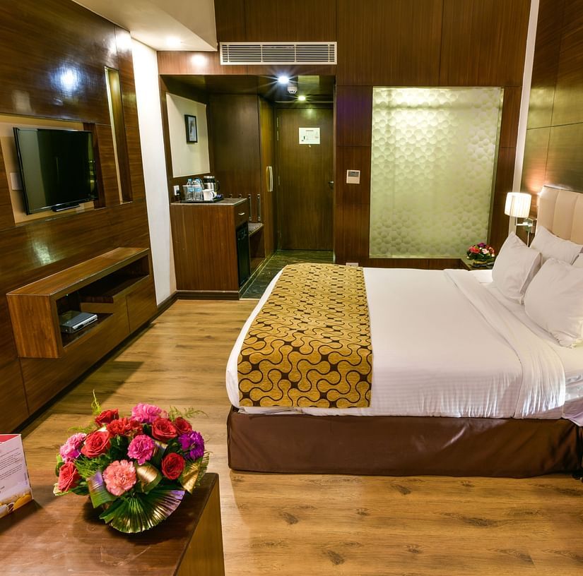 A well-appointed hotel room with wooden accents features a bed with multiple pillows, bedside tables with lamps and flowers, a television, and a glimpse into an adjoining area - Renest Jaipur