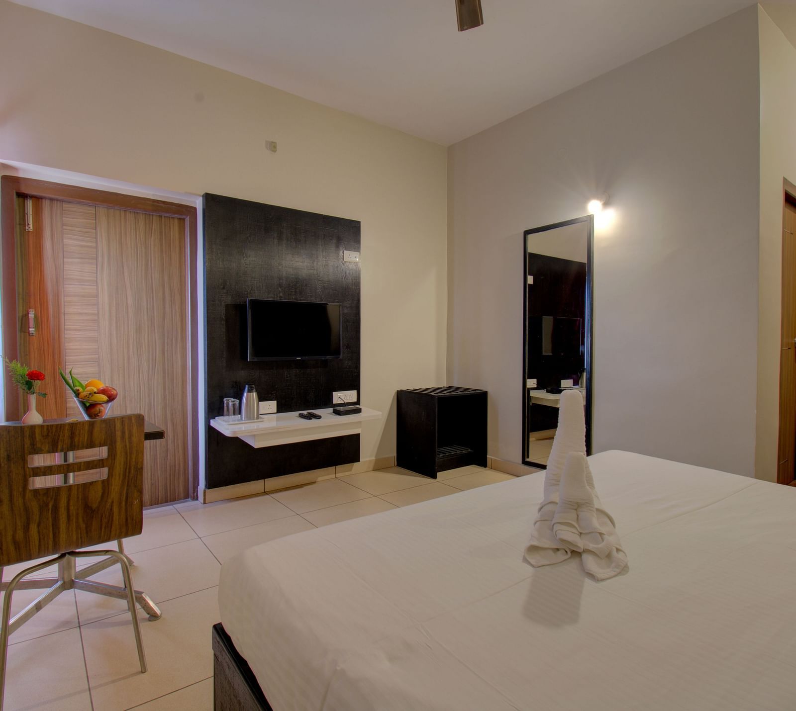 A bedroom with a bed, chair, TV unit, curtained windows, mirror and wooden door | Superior Room | woodstock resorts, Coorg