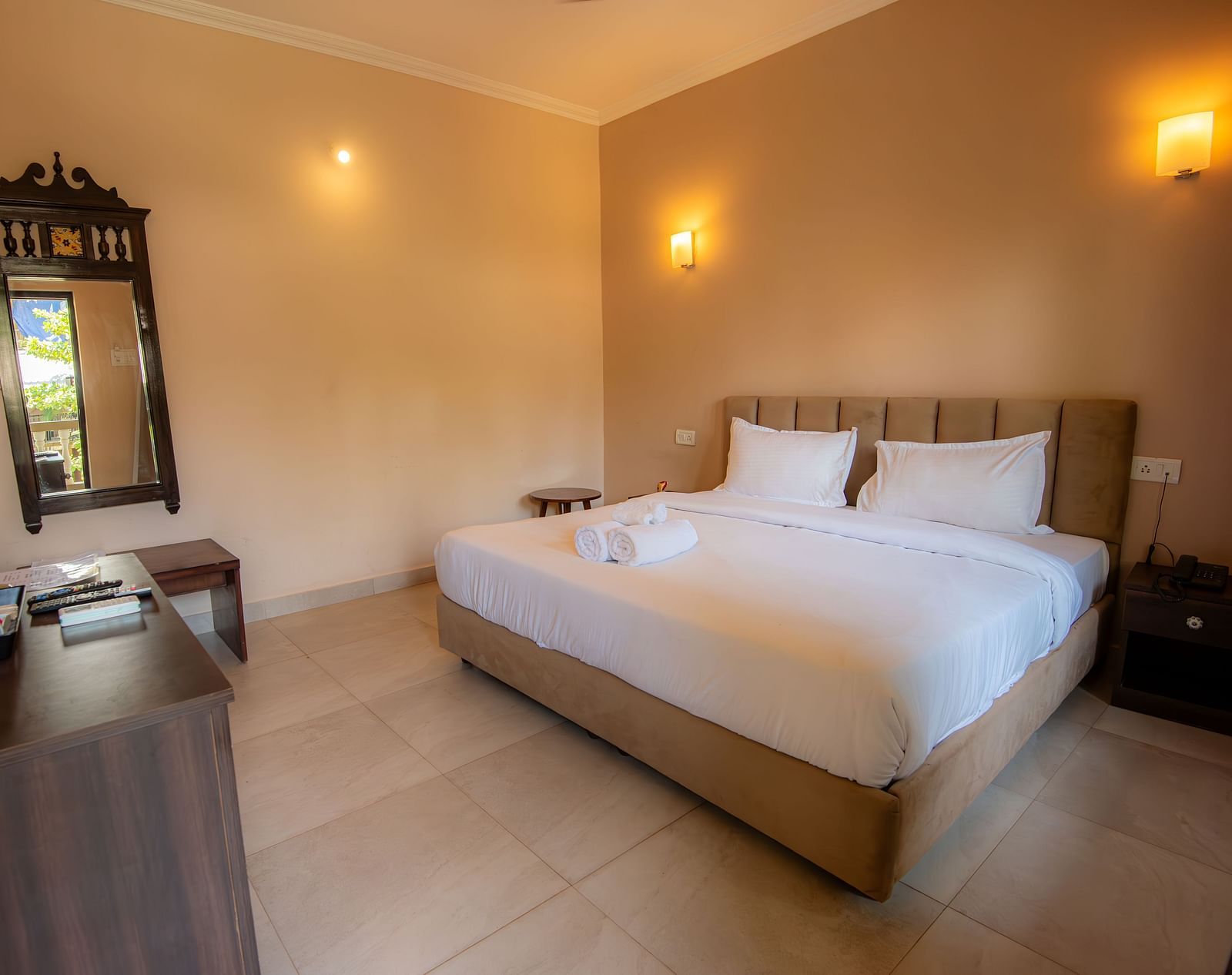 Well-lit bedroom with a large bed, bedside tables, and a decorative mirror placed on the wall at De Soul Santé, Morjim.