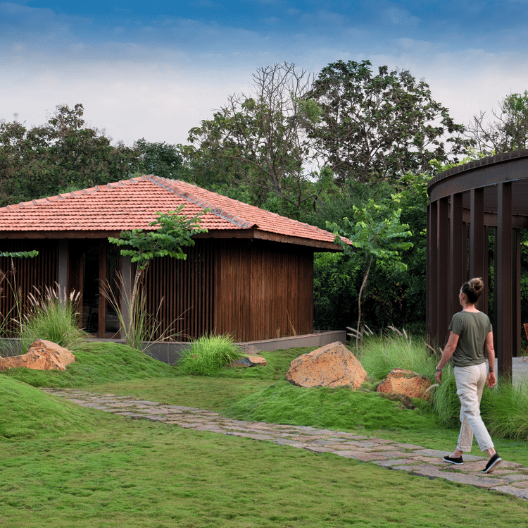 A person walking along a stone pathway surrounded by lush landscaped gardens at Aramness, with a wooden villa featuring a terracotta-tiled roof and a modern glass-walled pavilion nestled amid greenery and trees in the background.