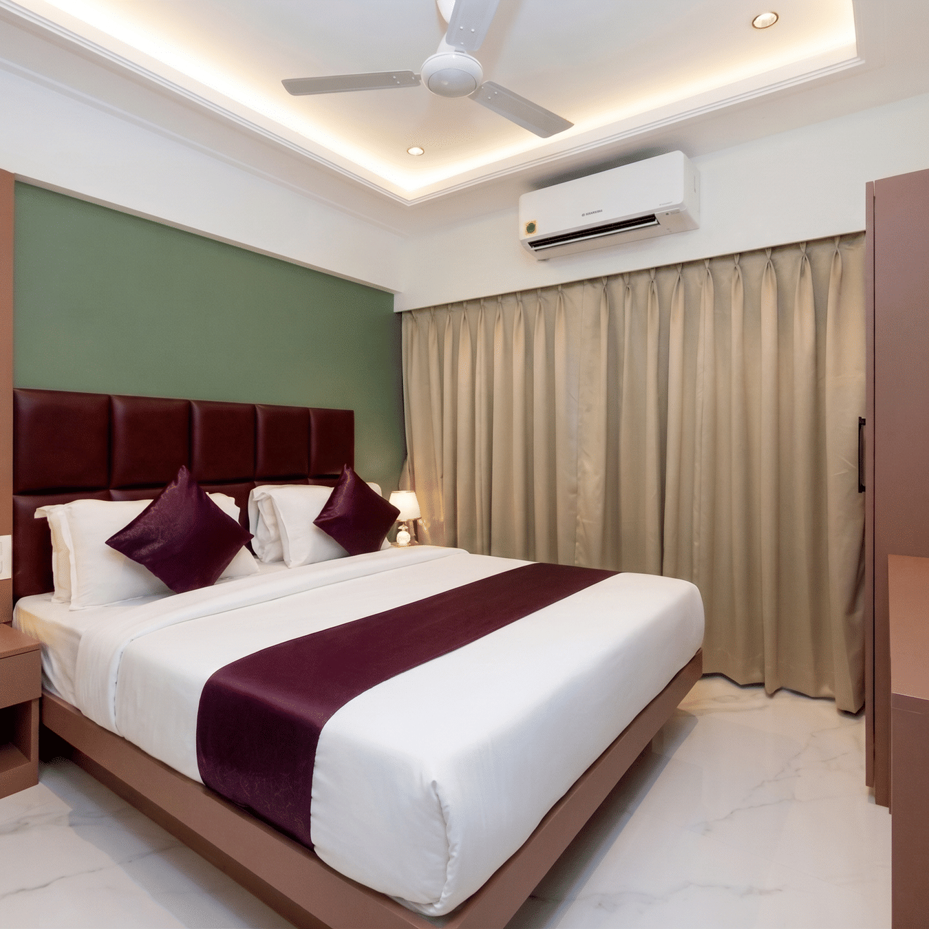 Deluxe Room featuring a king size bed with dark cushions, nightstand with a lamp, wall-mounted TV, A/C unit, and beige curtains at Home2 Residency, Mumbai Airport.