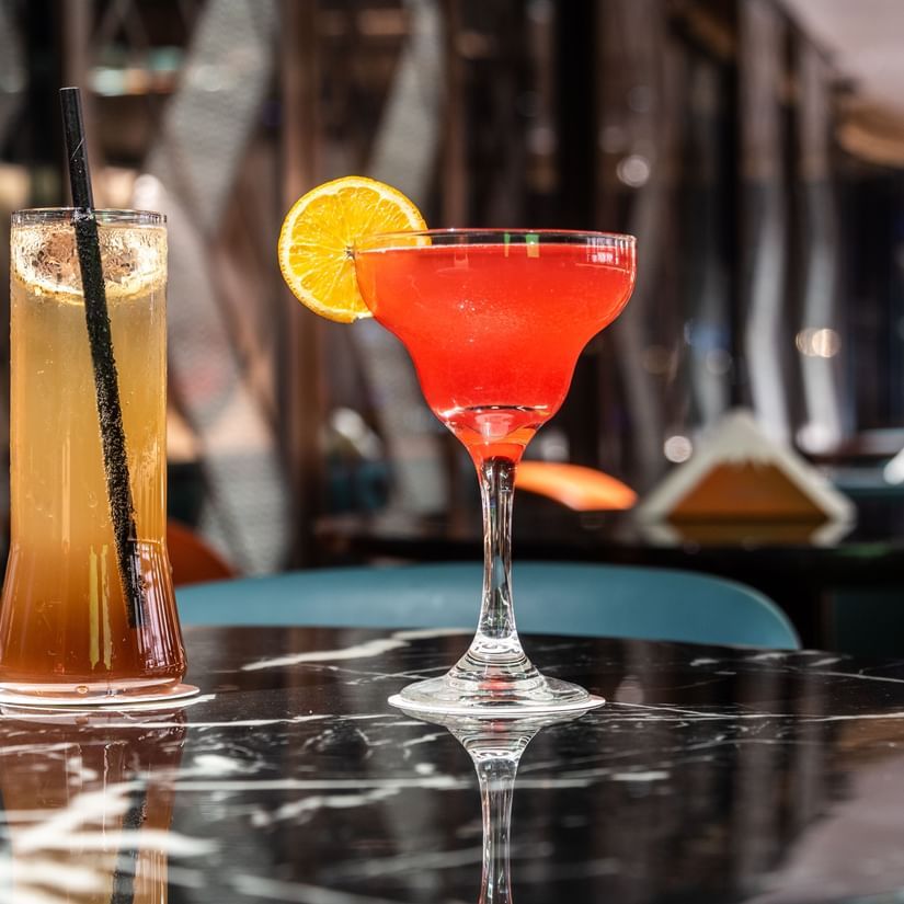 Close-up of a cocktail glass with a vibrant red drink garnished with orange and cherry, set on a reflective surface at Asiatic Lounge.