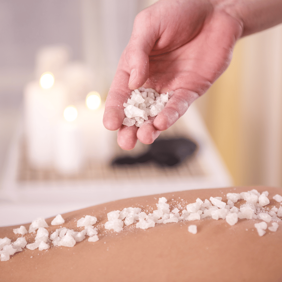 Close-up of therapist applying coarse crystals for body exfoliation on a client's back in a spa setting.