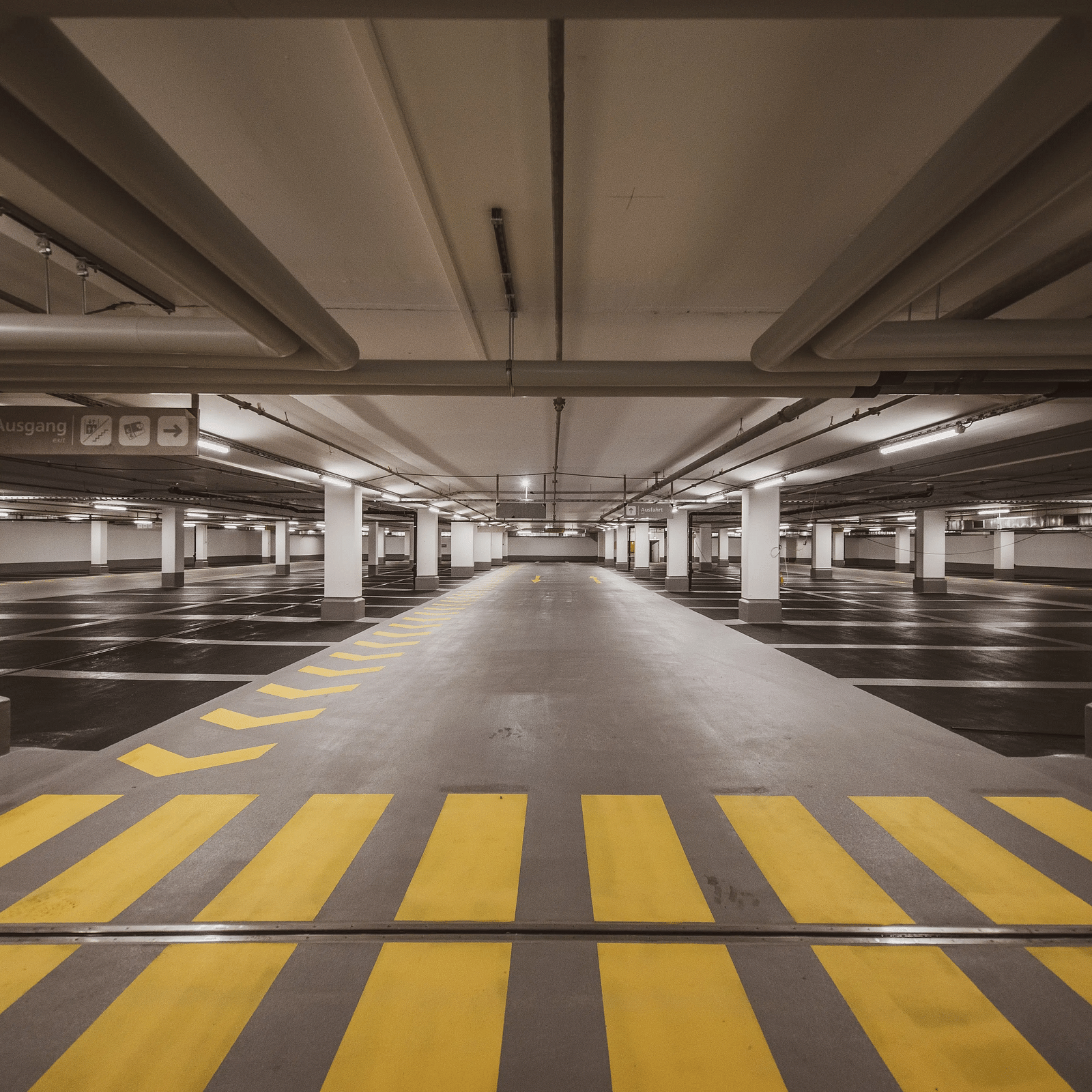 An empty underground parking garage with bright yellow pedestrian crossing lines, illuminated by ceiling lights and surrounded by rows of parking spaces