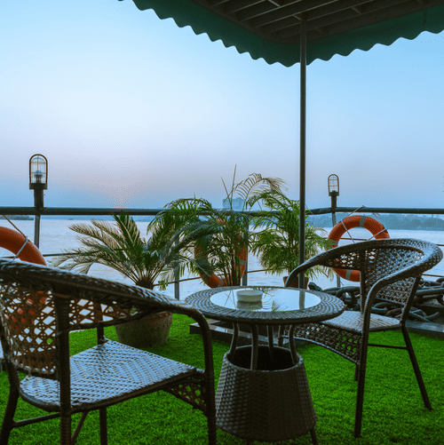 Relaxing outdoor balcony setting with rattan chairs and a table, overlooking a serene waterfront at dusk, surrounded by lush potted plants. - Polo Floatel Kolkata