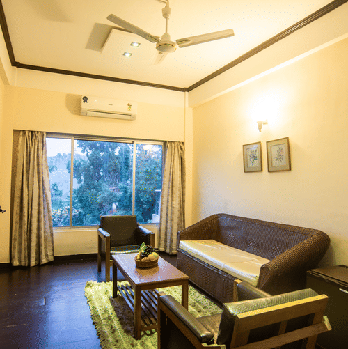 A cosy sitting room at Polo Tura Hotel featuring wicker and wooden furniture, a rug, large windows, and warm ambient lighting.