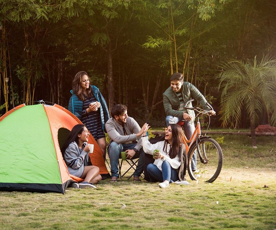 alt-text  A group of six people, including adults and teenagers, are gathered around a campsite with a green and orange tent. They are enjoying a meal and drinks together, with bicycles parked nearby.