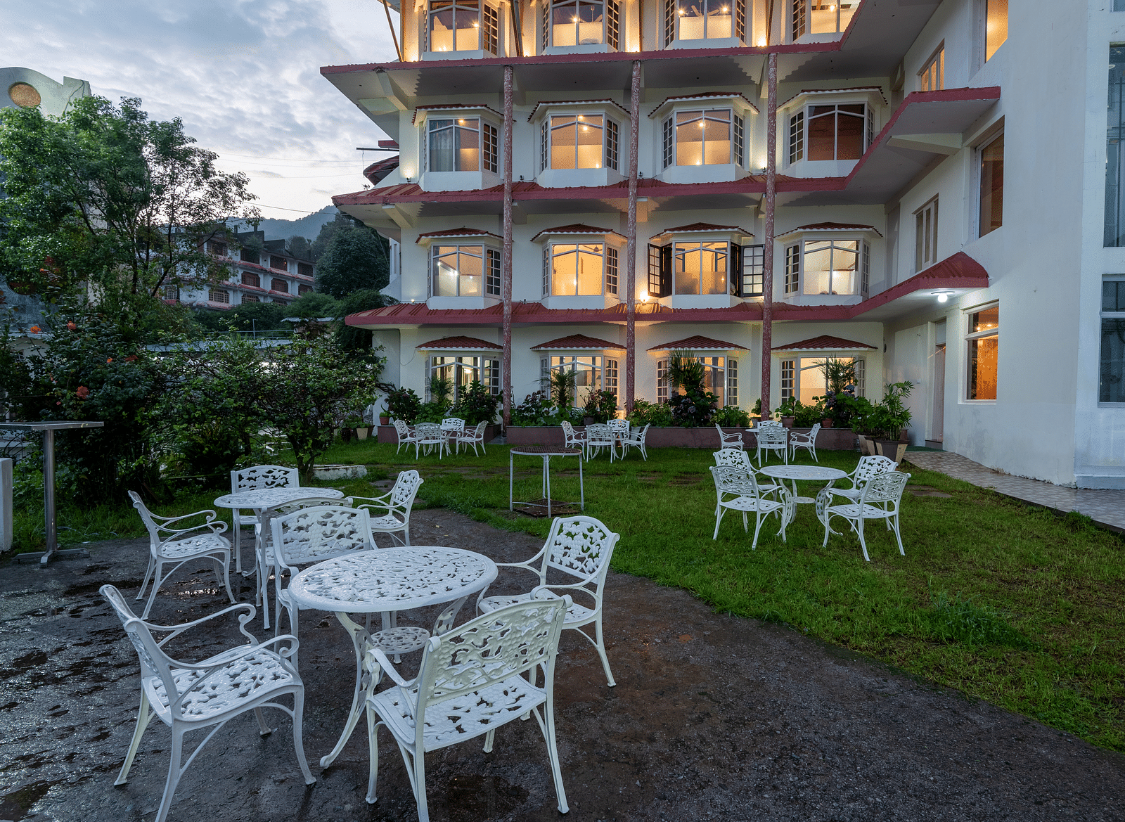 A resort garden with white outdoor tables and chairs, set against a building with lit windows. The area is surrounded by greenery and plants - DLS Meghavan Resort, Dharamshala